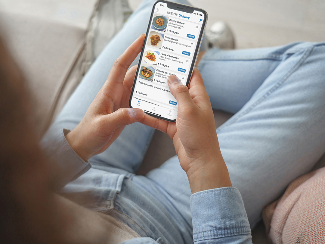 Una ragazza seduta su un divano sta consultando dal suo smartphone l'app Keep Up Delivery per ordini di cibo a domicilio