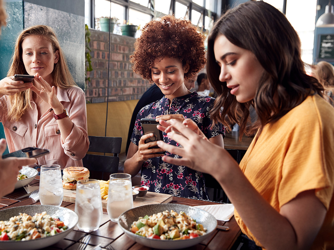 Tre ragazze sorridono mentre usano i loro smartphone sedute al tavolo di un ristorante